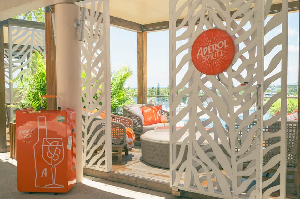 Covered terrace lounge with white geometric lattice screens, Aperol Spritz branded decor, and bay views.