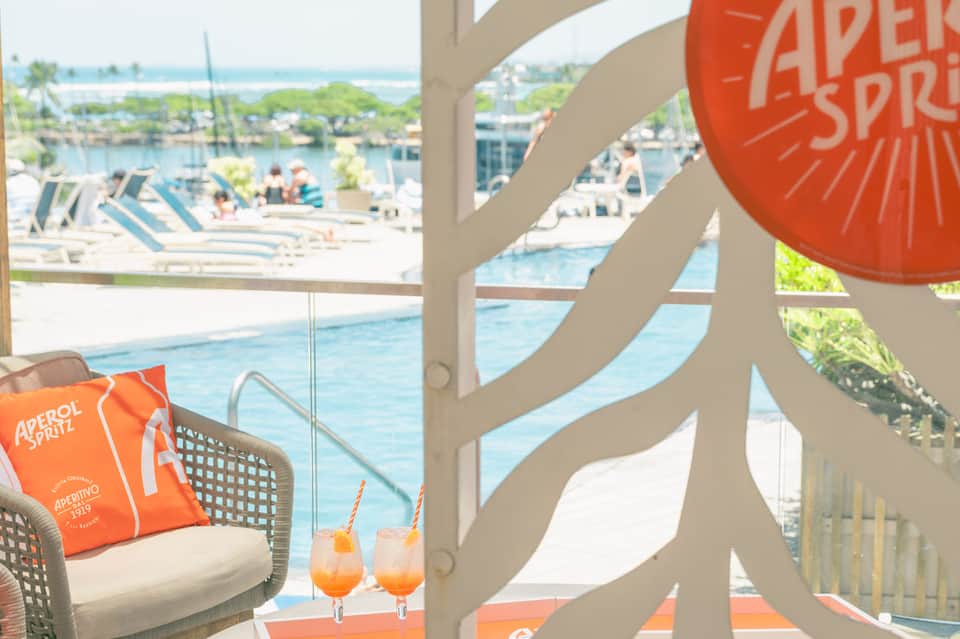 Waterfront lounge area with orange Aperol Spritz branding, cocktails, and pool view