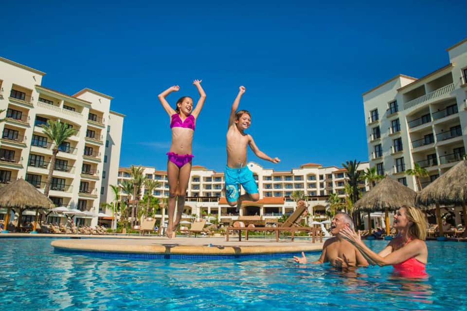 Children jumping into resort pool with parents watching, surrounded by palm trees and multi-story hotel buildings