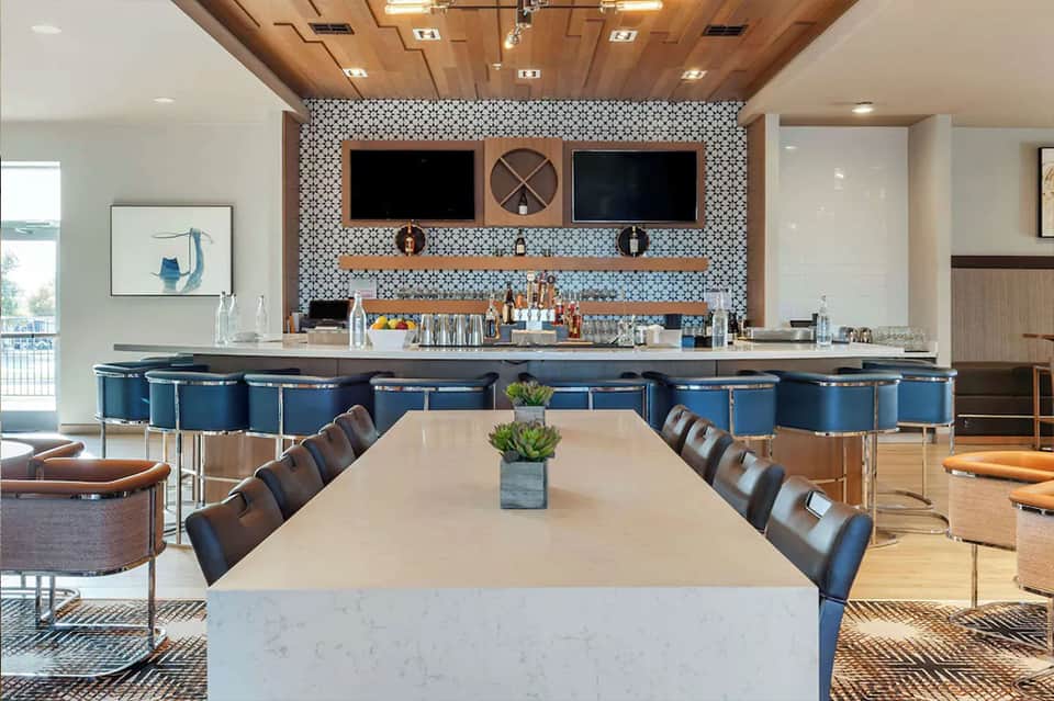 Modern hotel bar with blue stools, geometric tile backsplash, and dual TV screens above wooden shelving