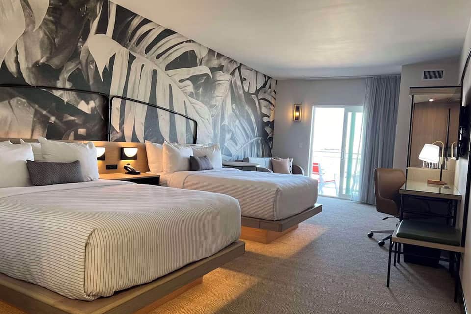 Modern hotel bedroom with two queen beds, striped bedding, botanical wall mural, and balcony access