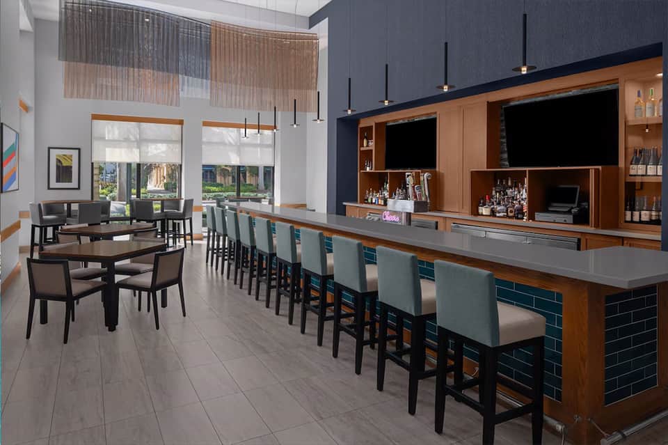 Modern hotel bar and lounge with wooden shelving, pendant lights, and high seating along a gray countertop