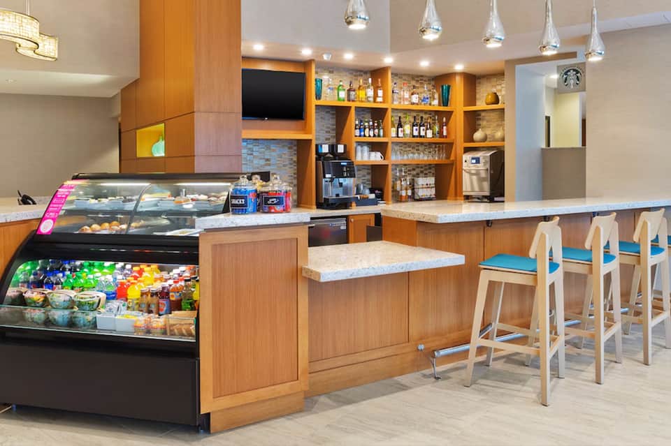 Modern hotel café with wooden shelving, pastry display case, Starbucks coffee station, and bar seating with blue cushioned chairs