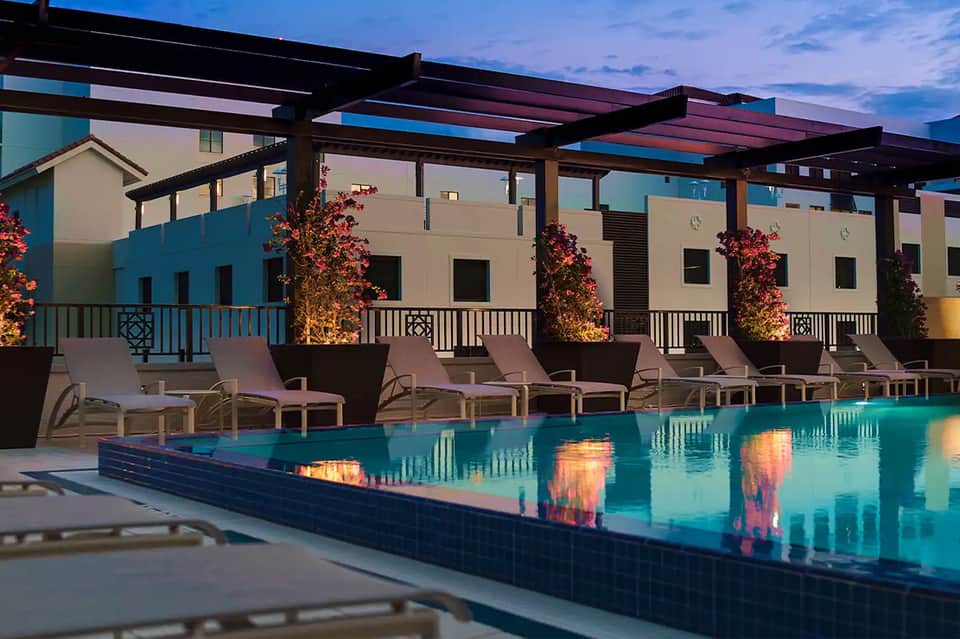 Contemporary pool deck at dusk with lounge chairs, pergola, and illuminated building facade