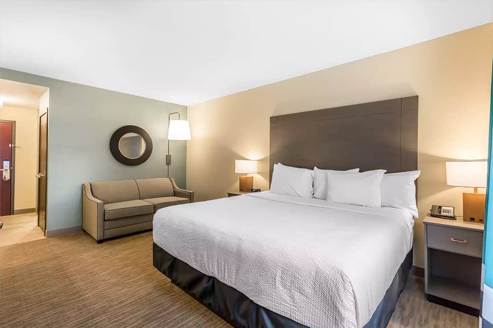 Hotel bedroom with queen bed, gray headboard, beige sofa, and warm lighting