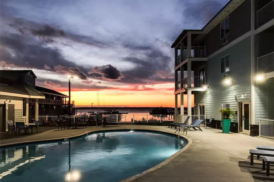 Lit swimming pool at dusk with waterfront view and dramatic sunset sky over buildings