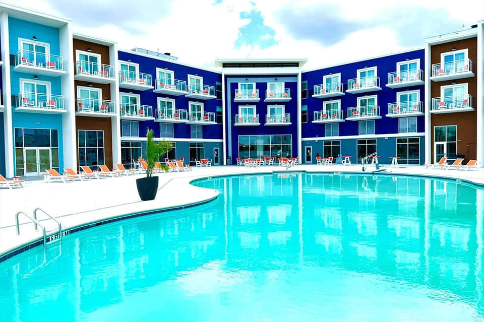 Resort swimming pool with bright turquoise water, white deck chairs, and modern blue and brown building facade.