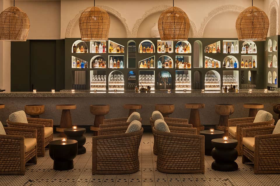 Upscale bar lounge with woven pendant lights, wicker seating, dark shelving with liquor bottles, and ambient candlelit tables