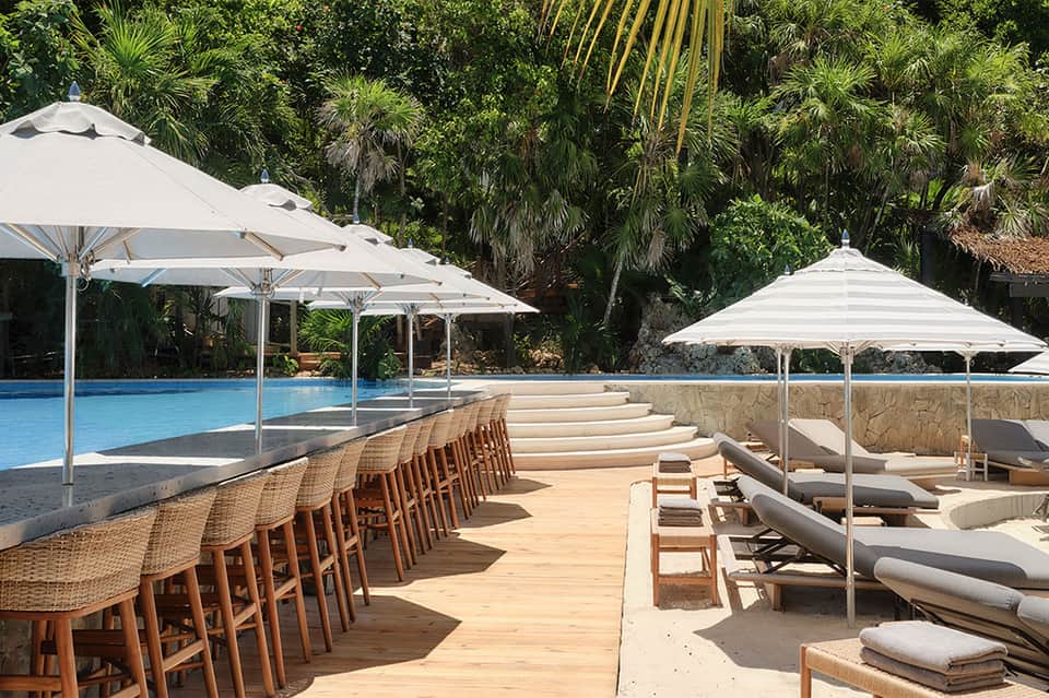 Resort pool deck with woven bar seating, white umbrellas, and loungers surrounded by tropical vegetation