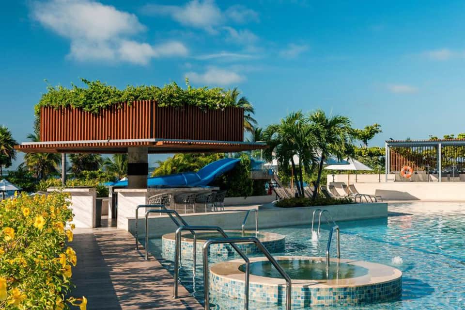 Resort pool area with water slide, hot tubs, and tropical landscaping under blue sky