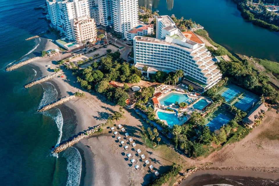 Aerial view of beachfront resort with white building, pools, and palm trees on turquoise ocean