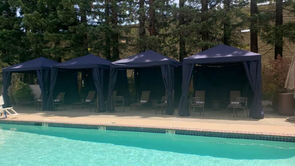 Three dark blue gazebos with curtains beside a bright turquoise swimming pool surrounded by tall pine trees