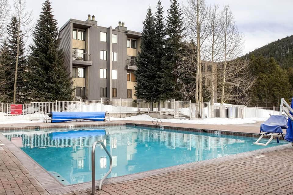Outdoor swimming pool in winter with snow, modern apartment building, and evergreen trees