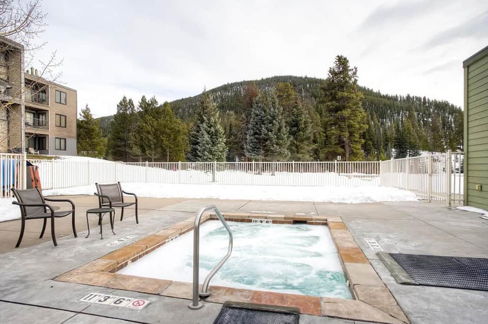 Mountain resort hot tub on snowy patio surrounded by forest, with lounge chairs and white railings