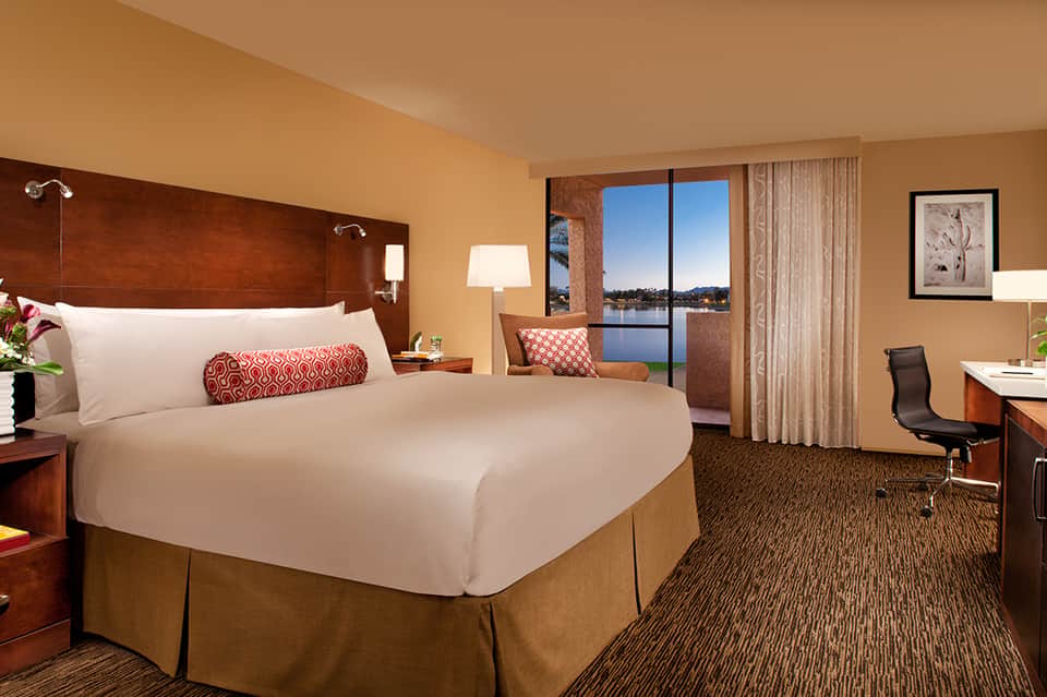 Luxury hotel bedroom with waterfront view, dark wood headboard, white bedding, and sitting area by floor-to-ceiling windows
