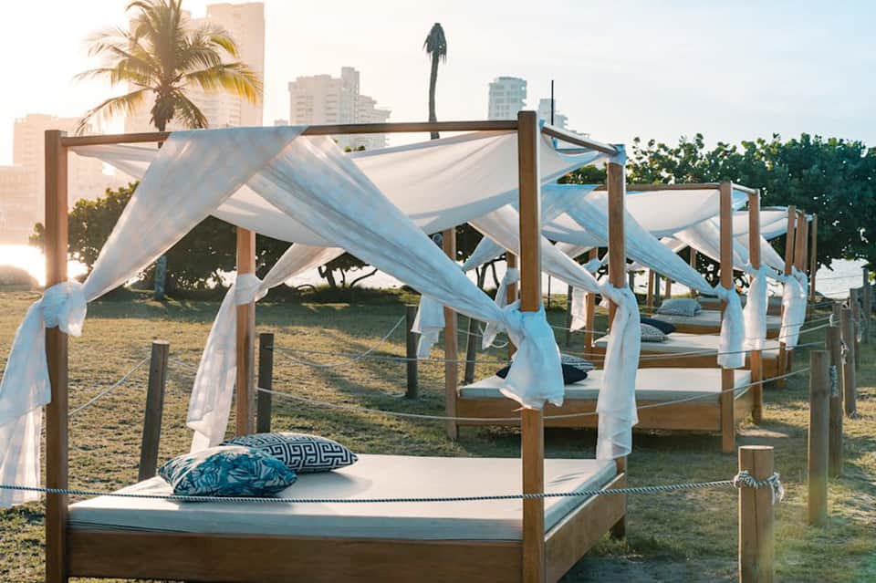 Beach cabana bed with white sheer curtains and blue pillows at sunset with city skyline backdrop
