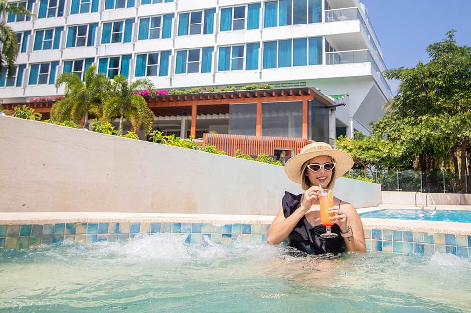 Woman in a pool at a modern resort holding a tropical drink, wearing a hat and sunglasses