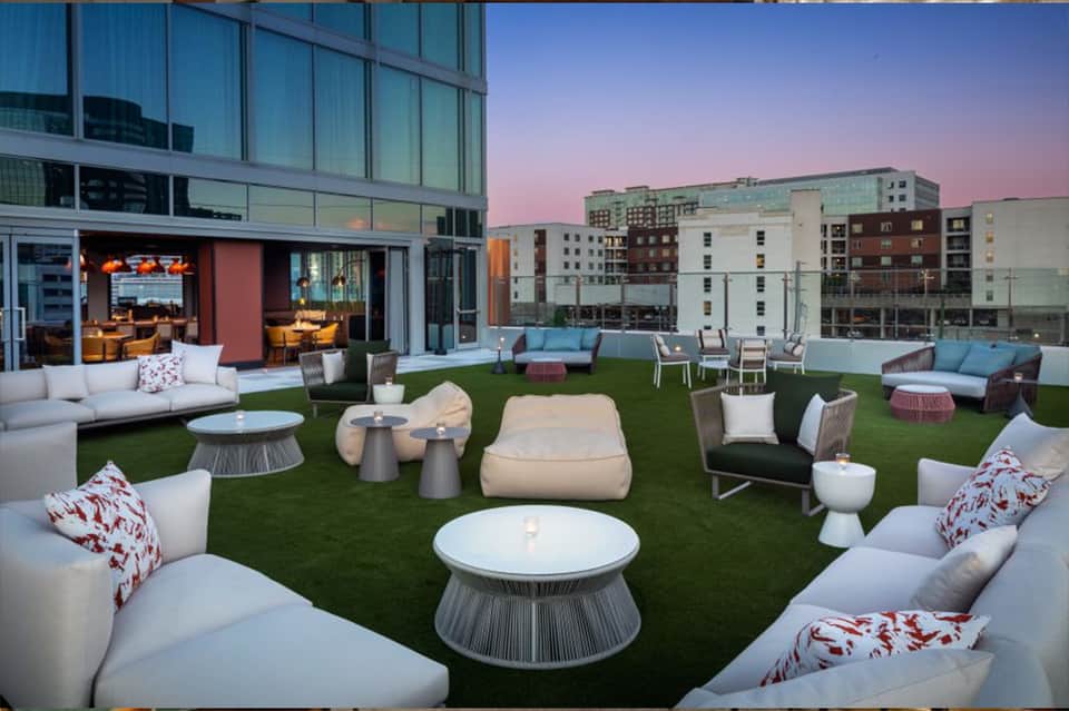 Rooftop lounge with artificial turf, modern seating clusters, fire pits, and interior space overlooking city skyline at dusk