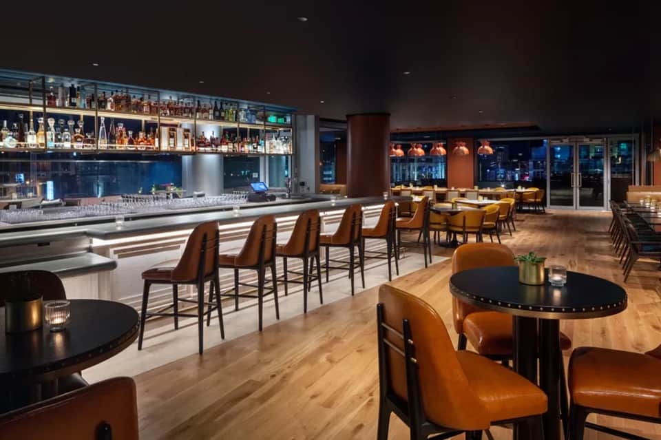 Upscale bar lounge with illuminated liquor shelves, cognac-colored leather seating, black tables, and city night views