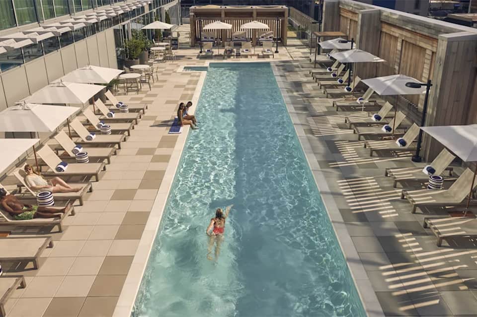 Rooftop pool deck with lap pool, lounge chairs with white umbrellas, and surrounding city skyline views