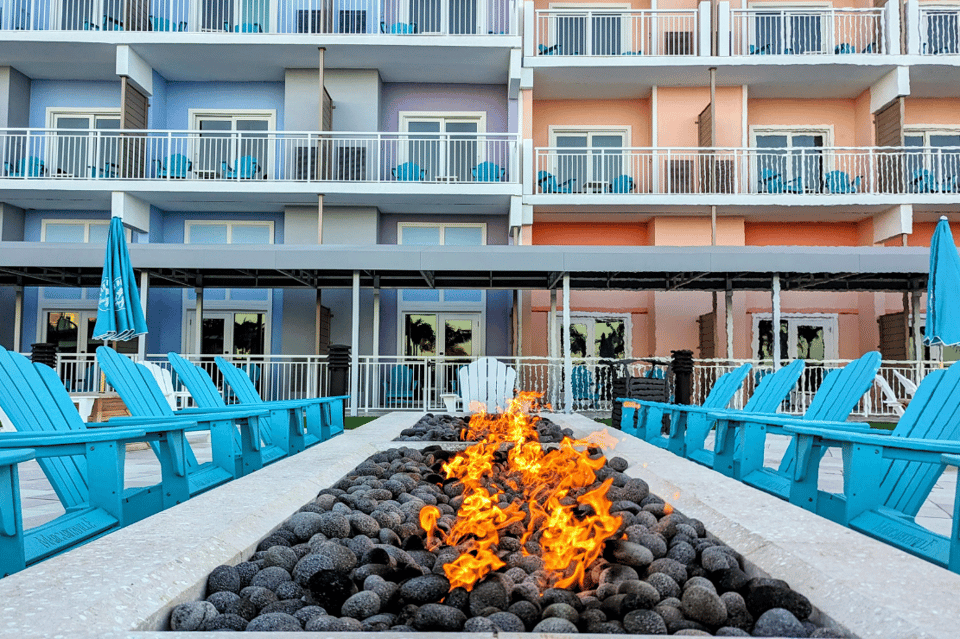 Beachfront fire pit surrounded by bright blue Adirondack chairs with colorful hotel buildings in background