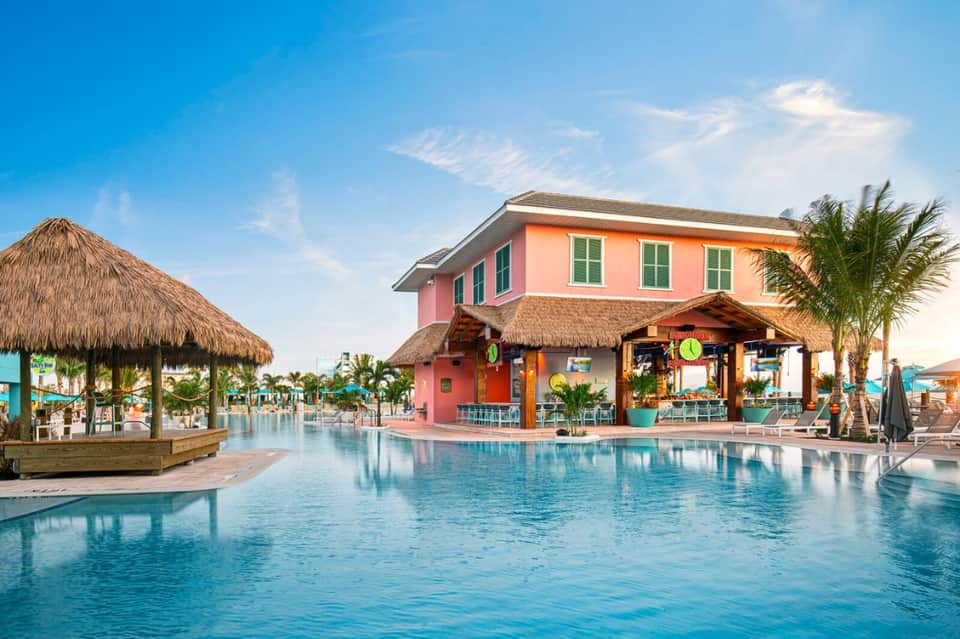 Tropical resort pool with pink main building, thatched-roof cabanas, and palm trees under blue sky