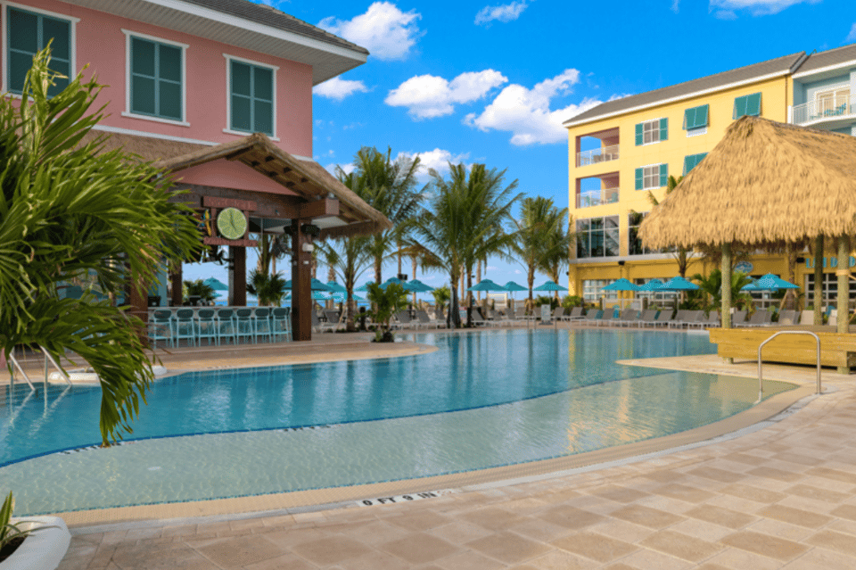 Resort pool area with colorful buildings, tiki bar, palm trees, and beach access visible
