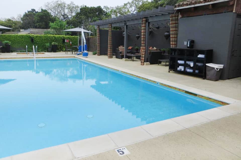 Resort swimming pool with concrete deck, cabanas, umbrellas, and poolside amenities surrounded by hedges