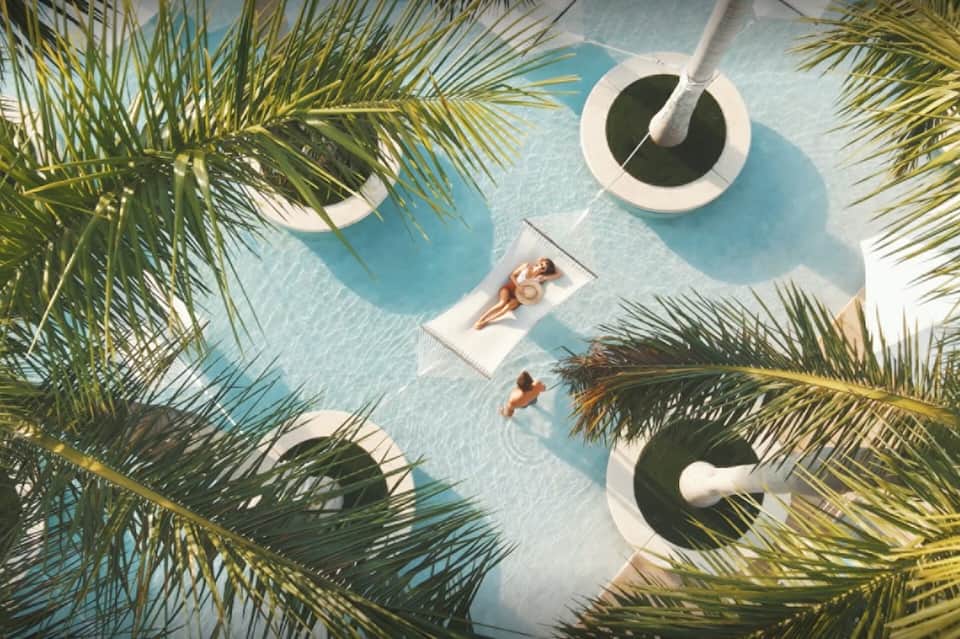 Aerial view of light blue pool with palm trees, white float raft, and sunbathing guests