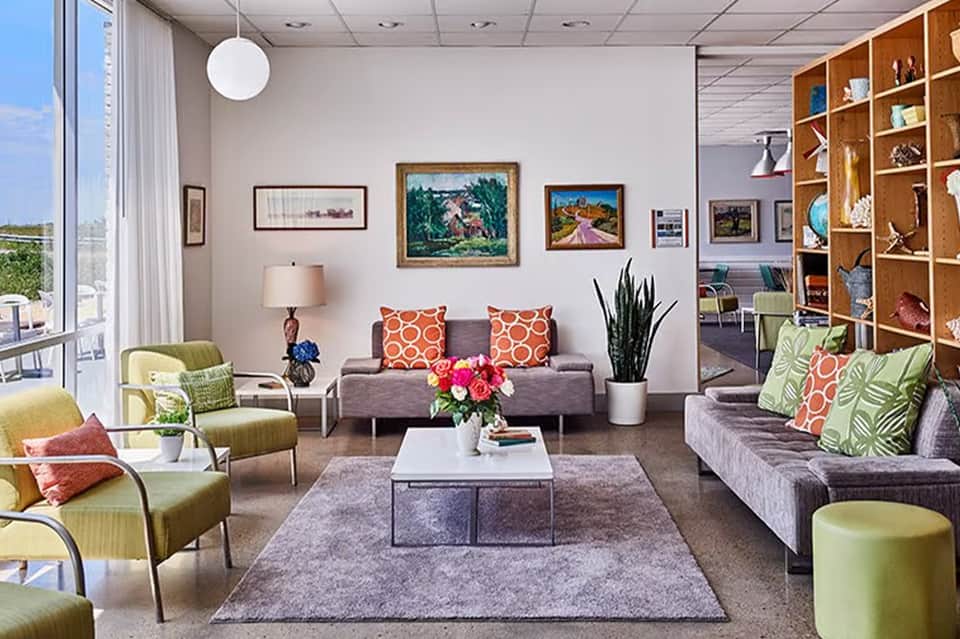 Bright lounge area with colorful pillows, artwork, and wood shelving near large windows