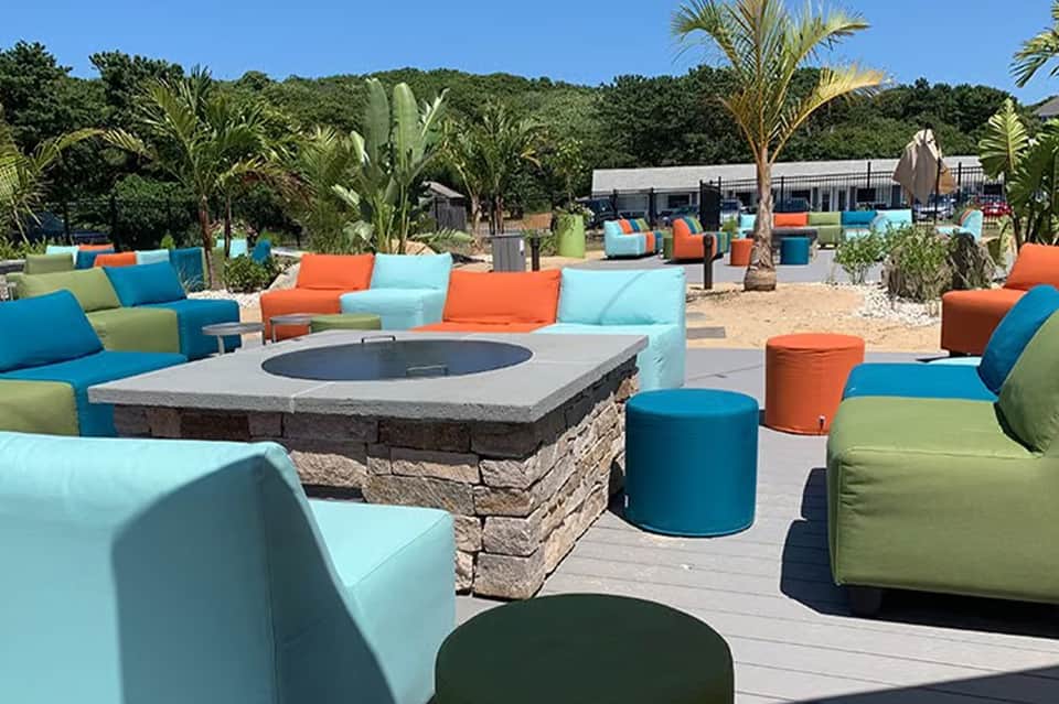 Colorful outdoor seating area with blue, orange, and lime cushioned furniture surrounding a stone fire pit on sandy ground