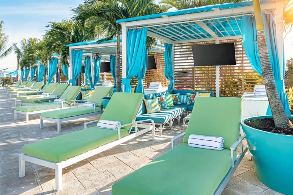 Beachside cabanas with turquoise pergolas, green loungers, and palm trees under clear sky