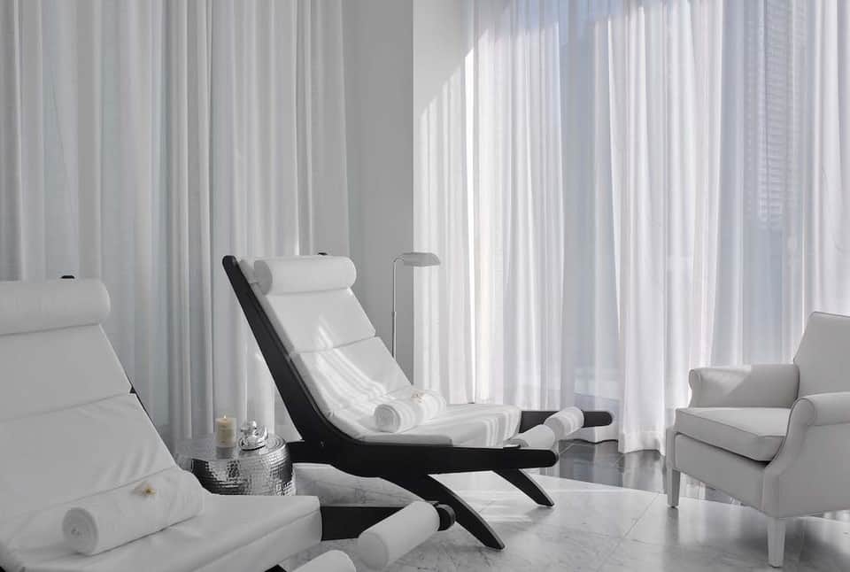 Modern spa relaxation room with white loungers, black accent chair, sheer curtains, and natural light