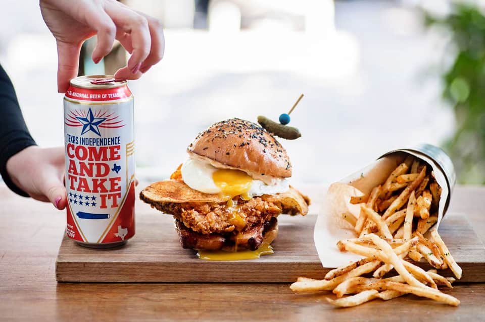 Fried chicken sandwich with egg and cheese, fries, and Texas Independence beer on wooden board
