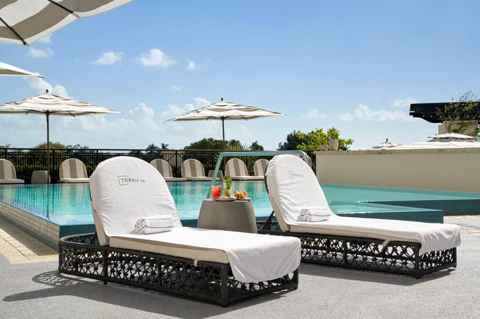 Two lounge chairs with white cushions beside turquoise pool under umbrellas