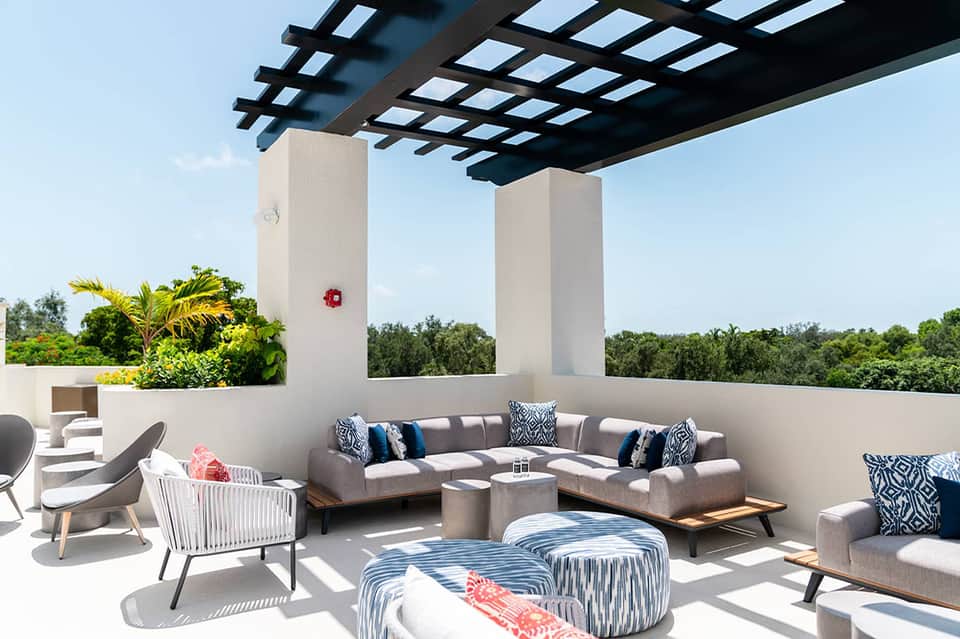 Modern rooftop terrace with gray sectional seating, patterned pillows, pergola shade structure, and tropical plants