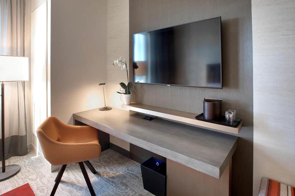 Modern hotel room desk with wall-mounted TV, desk lamp, orchid plant, and contemporary furnishings in neutral tones
