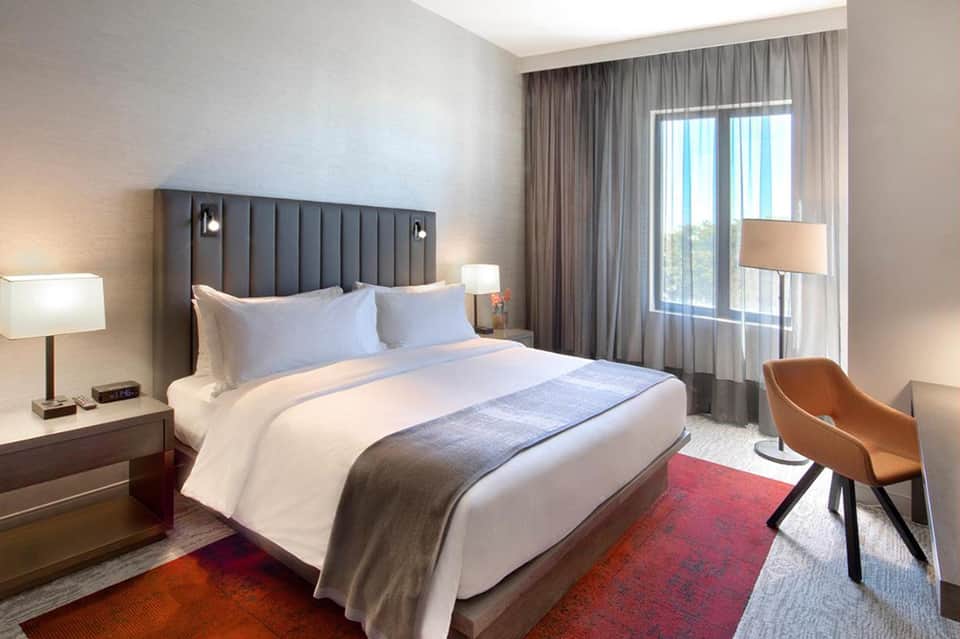 Modern hotel bedroom with gray upholstered headboard, white bedding, and ocean view through glass doors