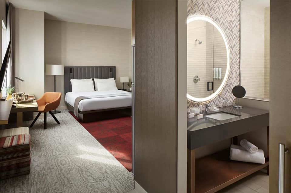 Modern hotel room with king bed, ensuite bathroom with illuminated round mirror, and contemporary furnishings