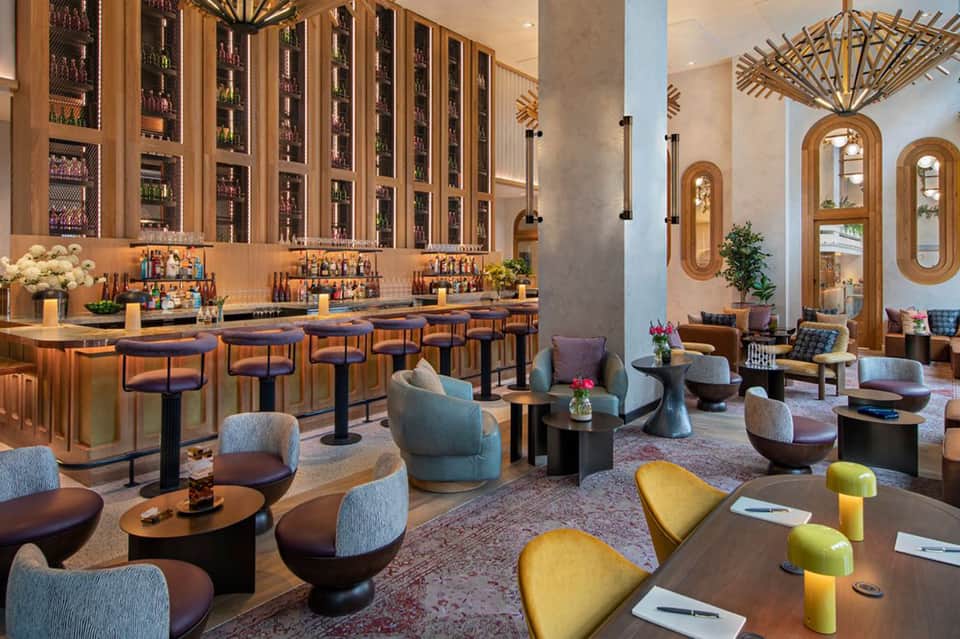 Upscale bar and lounge with wooden wine display, navy bar stools, colorful accent chairs, and architectural mirrors