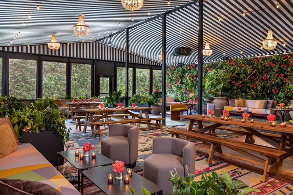Elegant rooftop lounge with black striped pergola, wooden furniture, flowering plants, and crystal chandeliers