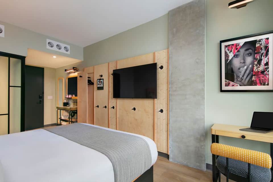 Modern hotel bedroom with plywood storage walls, flat-screen TV, desk with laptop, and contemporary furnishings