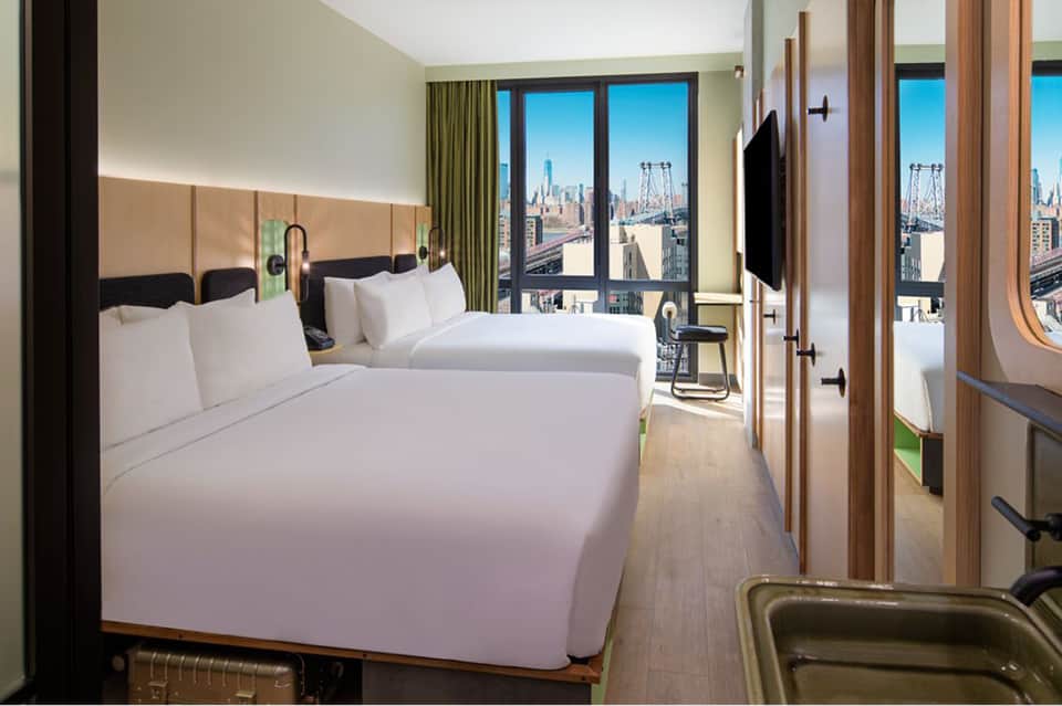 Luxury hotel bedroom with two queen beds, green curtains, and expansive windows overlooking city skyline