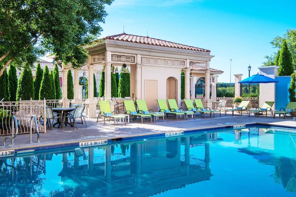 Resort pool with classical architecture, green lounge chairs, and palm trees under blue sky