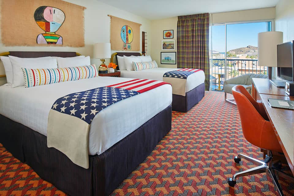 Hotel room with two queen beds, colorful American flag bedding, modern artwork, and balcony views of hills