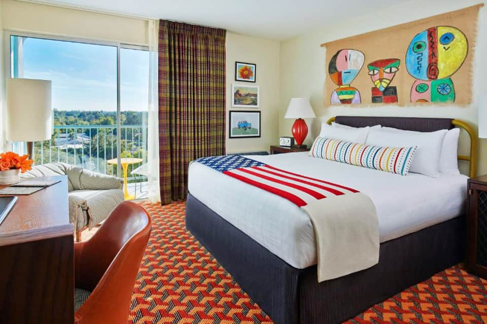Bright hotel bedroom with queen bed, ocean view balcony, colorful modern artwork, and striped bedding