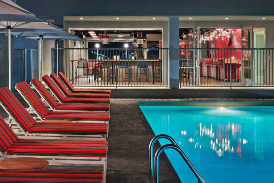 Resort outdoor pool at night with red lounge chairs, terrace seating, and illuminated bar area