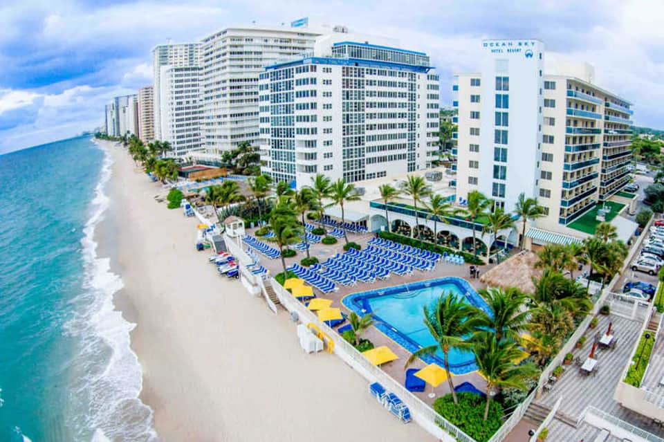 Beachfront resort with white multi-story buildings, blue lounge chairs on sandy beach, ocean waves, and palm trees