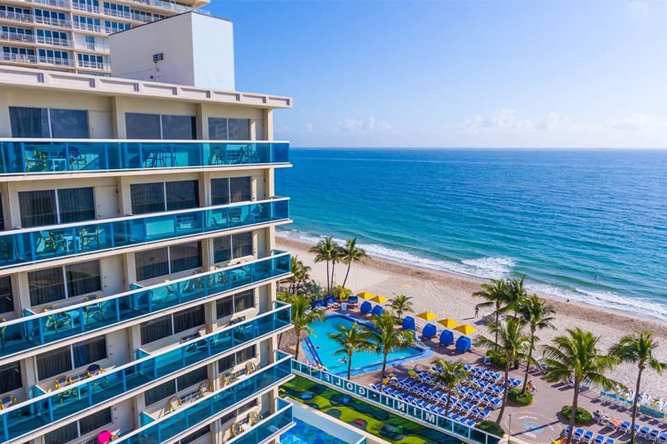 Beachfront resort with multi-story building, blue balconies, pool, palm trees, and sandy beach with ocean view