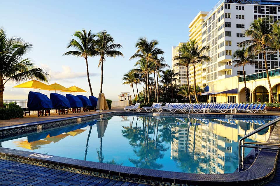 Beachfront pool with palm trees, yellow umbrellas, and high-rise hotel building with yellow facade